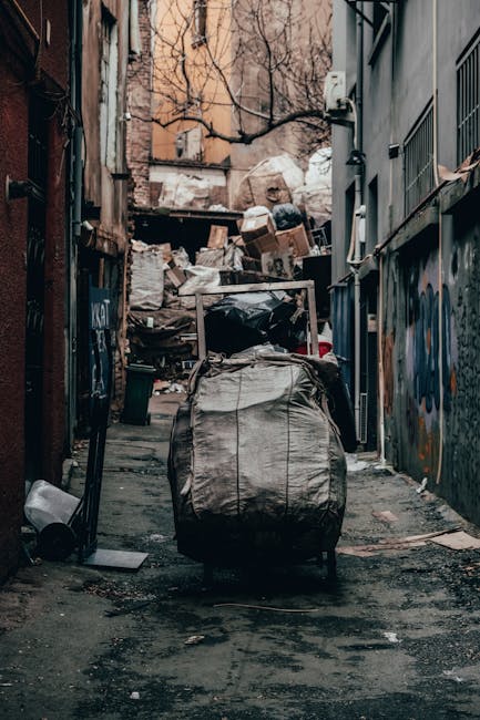 A large, rusted red metal skip positioned against a grey concrete wall on a paved urban street, with multiple black and white plastic rubbish bags stacked in front of it. The skip has visible signs of